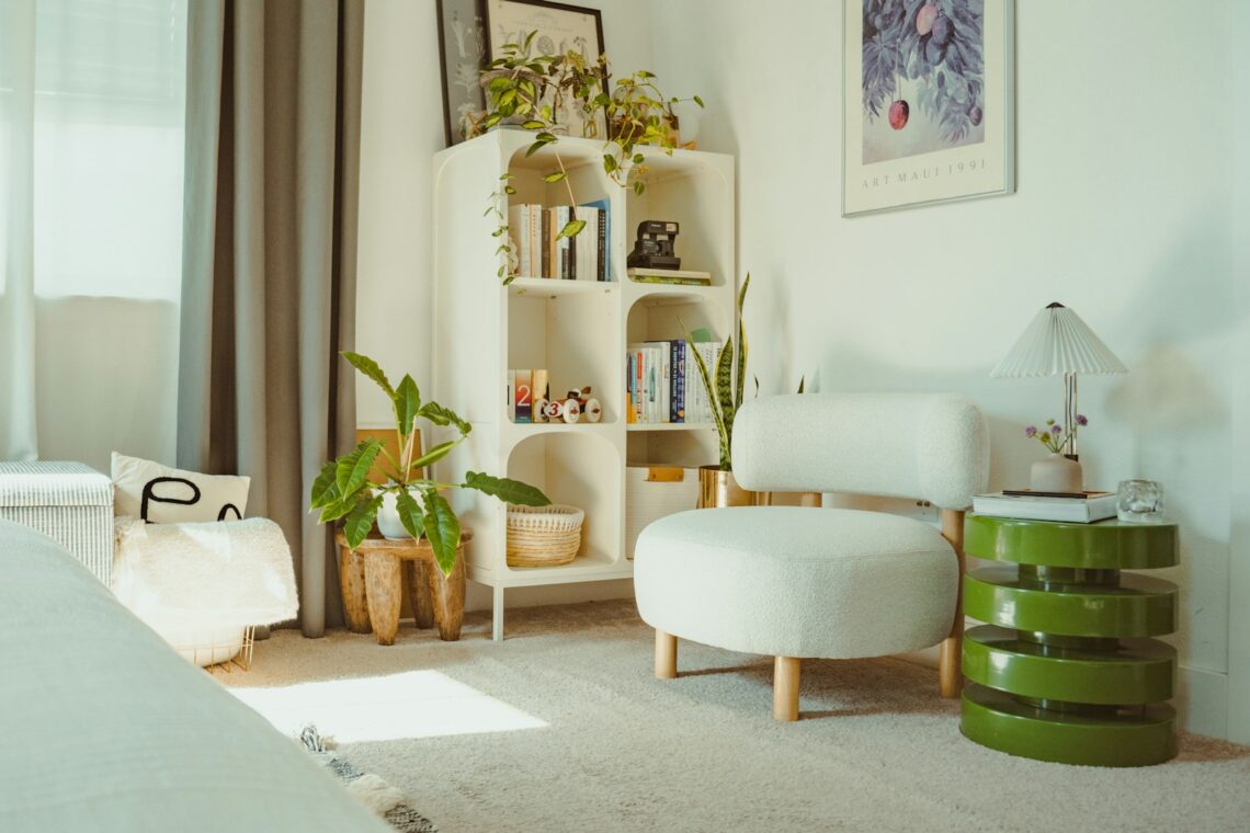 Bright cozy apartment room with white organic bookcase filled with books and plants, fluffy armchair nearby