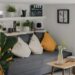 Cozy small studio apartment living room with wall shelves holding plants and gadgets, grey sofa with mustard yellow pillows