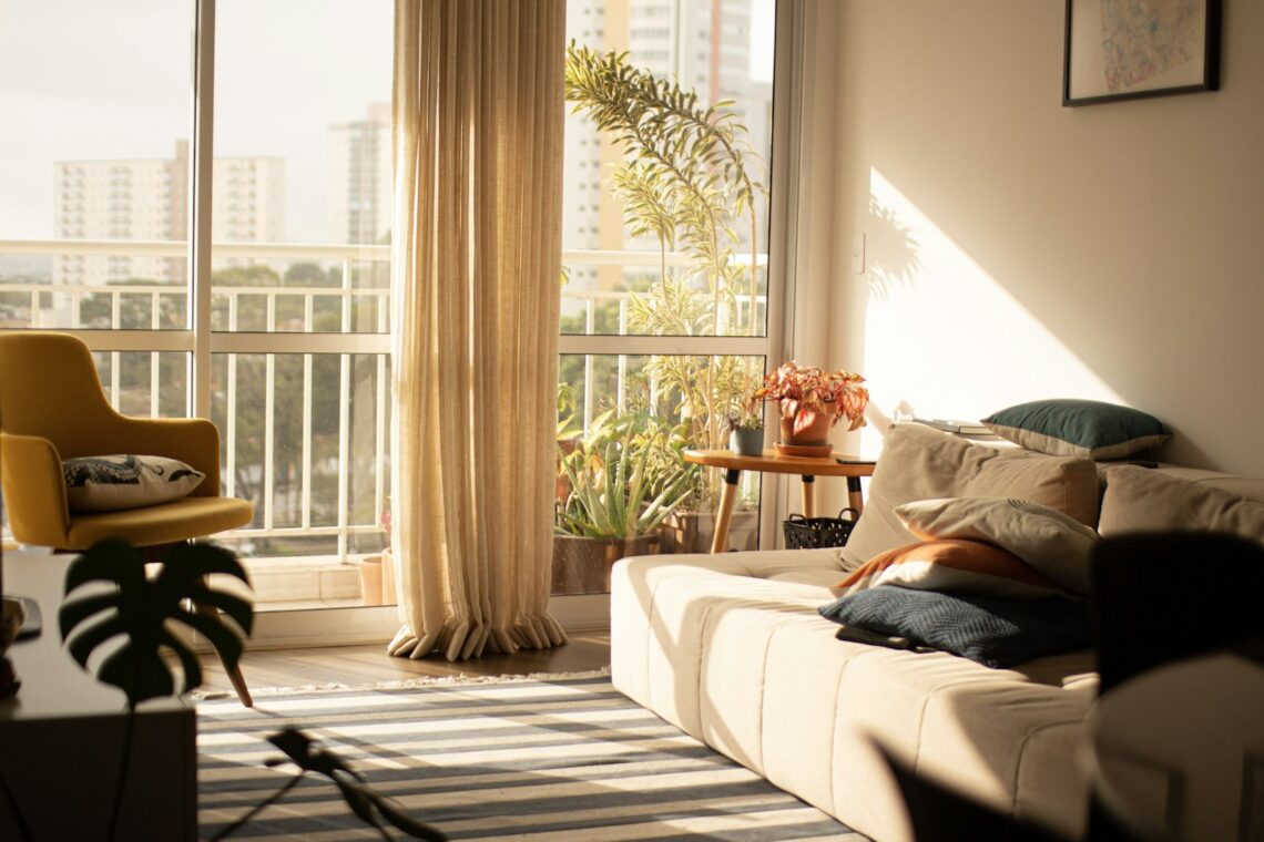 Sun-drenched small apartment living room with floor-to-ceiling windows, sofa, yellow armchair and plants, no TV