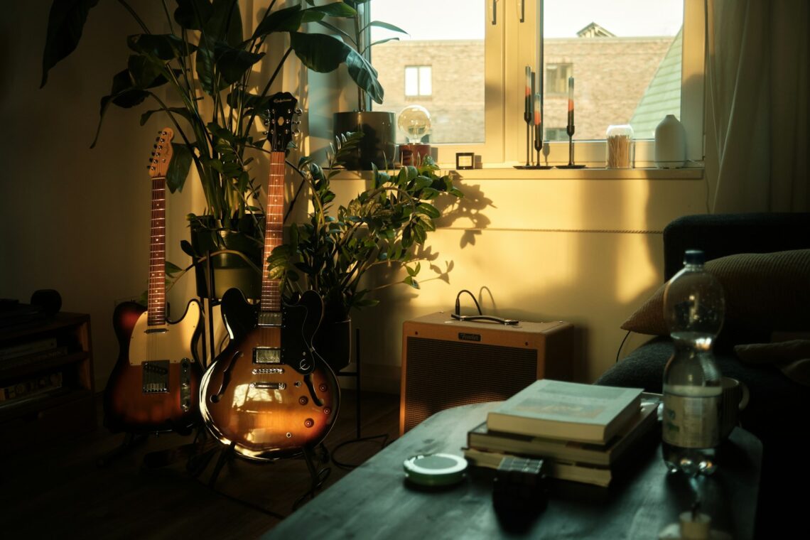 Moody apartment living room with warm golden light, guitars leaning against wall, stacked books and lush plants in a cozy renter space