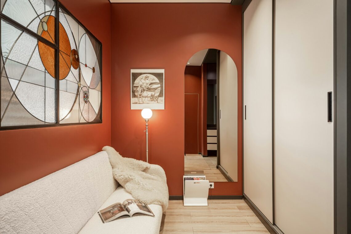 Cozy small apartment room with deep terracotta red walls, arched doorway, cream bouclé sofa, stained glass window, and warm floor lamp creating an intimate earthy atmosphere