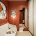 Cozy small apartment room with deep terracotta red walls, arched doorway, cream bouclé sofa, stained glass window, and warm floor lamp creating an intimate earthy atmosphere