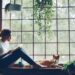 Woman reading a book on a window seat next to her dog, surrounded by hanging plants and natural light
