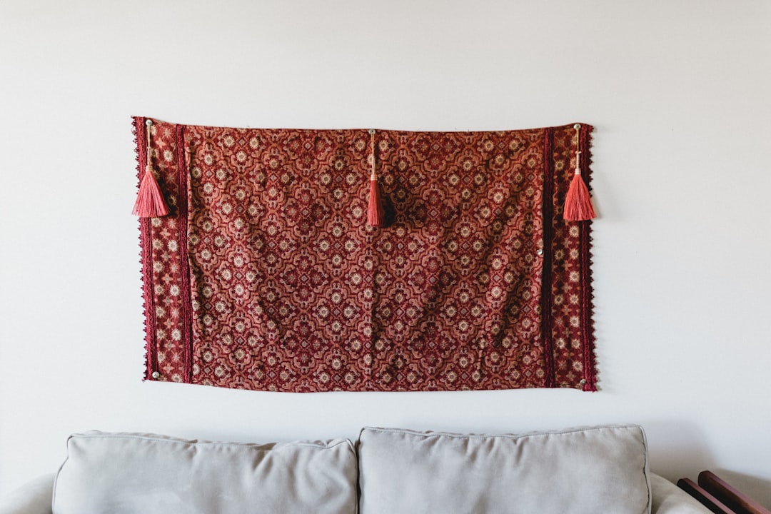 Red and rust patterned textile tapestry with pink tassels hung on a white wall above a light gray sofa
